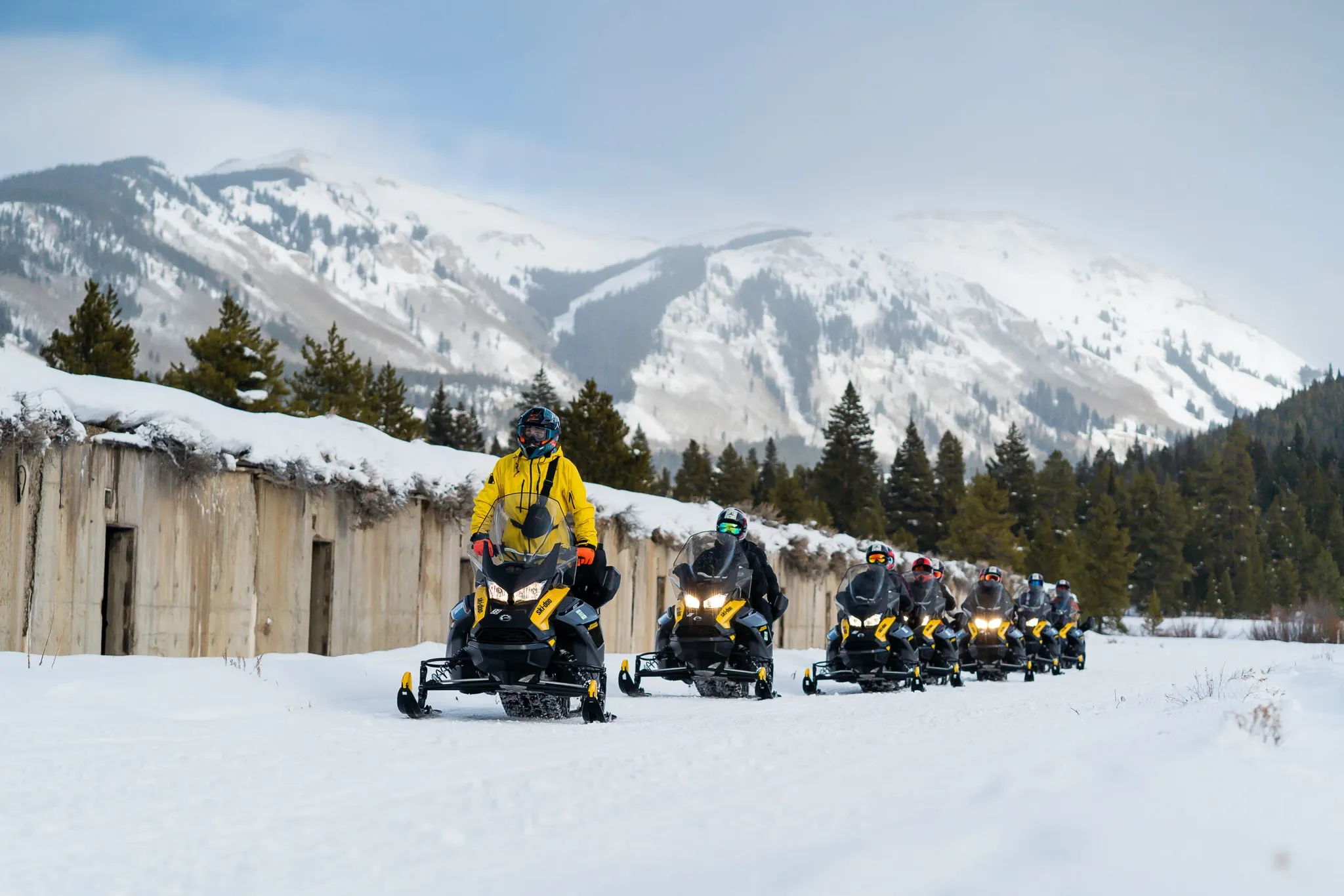 line of snowmobile riders next to flat top buildings with sweeping mountain views.
