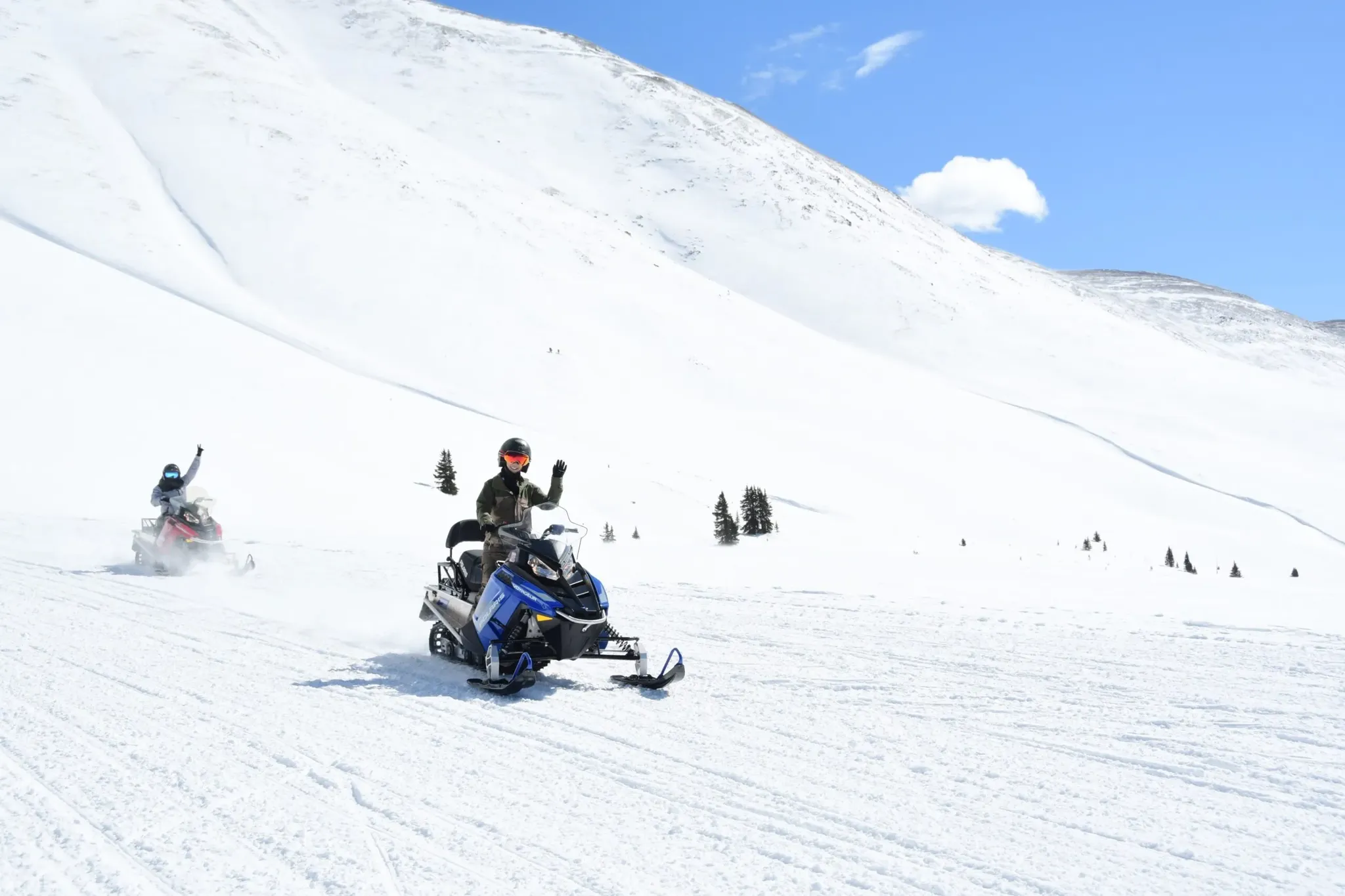 snowmobile riders waving from the side of a snow covered mountain.